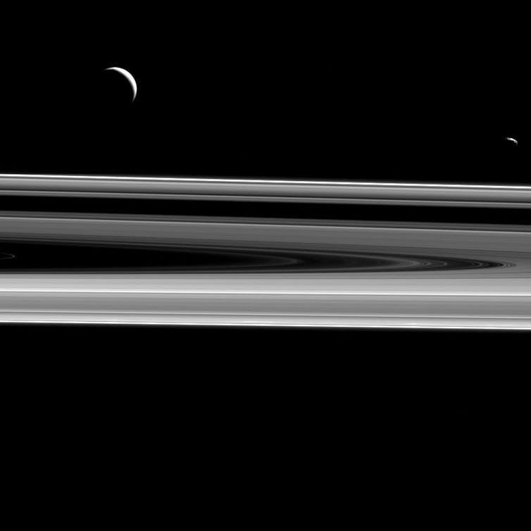 Uma imagem em escala de cinza com um fundo preto. Os anéis de Saturno estão na parte inferior da imagem e aparecem como uma meia-oval listrada em vários tons de cinza. Duas luas parcialmente eclipsadas pairam sobre os anéis, aparecendo como crescentes de luz.